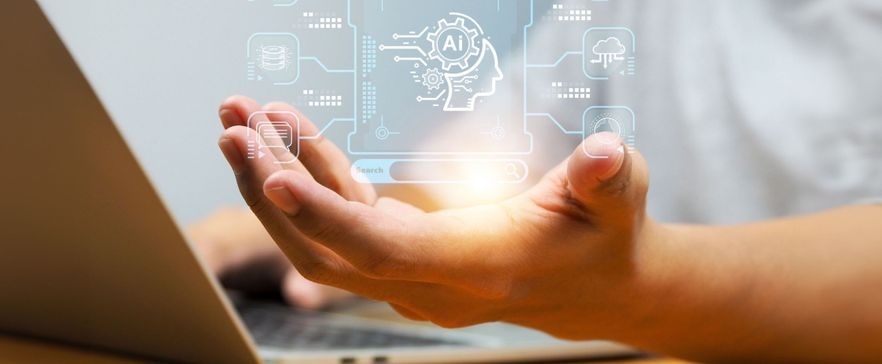 Eine Hand hält ein leuchtendes, illusorisches Bild, das aus technischen und digitalen Symbolen besteht. Darunter befinden sich Zahnräder und ein menschliches Profil, beschriftet mit "AI". Im Hintergrund ist ein Laptop zu erkennen.