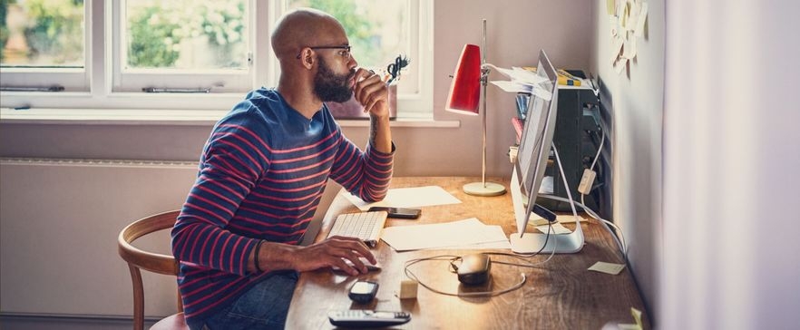 Das Bild zeigt einen Mann mit Brille, der an einem Schreibtisch sitzt und auf einen Computerbildschirm schaut. Er trägt ein gestreiftes Langarmshirt und hat eine nachdenkliche Haltung. Auf dem Schreibtisch befinden sich verschiedene Gegenstände.