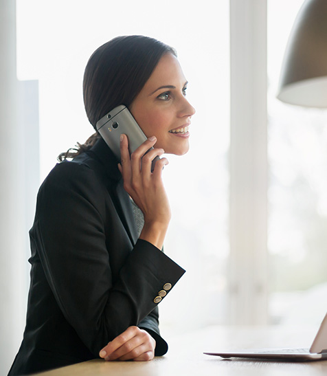 Frau mit Smartphone am Ohr sitzt am Laptop und macht telefonisch eine Vertragsverl&auml;ngerung.