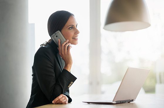 Frau mit Smartphone am Ohr sitzt am Laptop und macht telefonisch eine Vertragsverl&auml;ngerung.